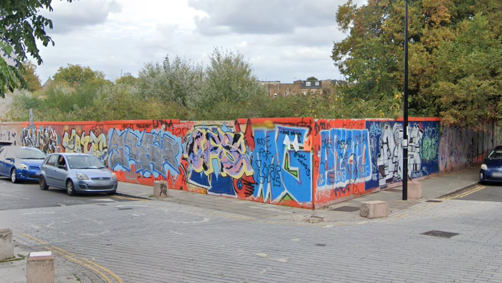 A street view image of orange hoarding around the waste ground site, which has graffiti sprayed on it. Cars can be seen parked on the roads around the site