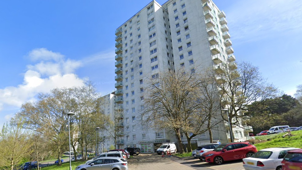 A tall apartment under clear blue skies. There is a small car park with a handful of cars parked in it. 