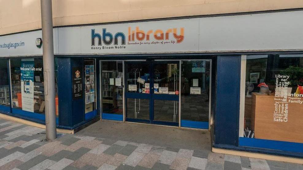 The exterior of the library, which is glass fronted and has white signage with hdn library written on it in blue and orange.