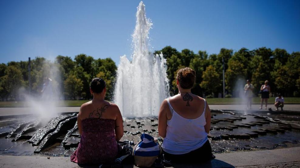Two women look at a fountain on a sunny day with their backs to the camera