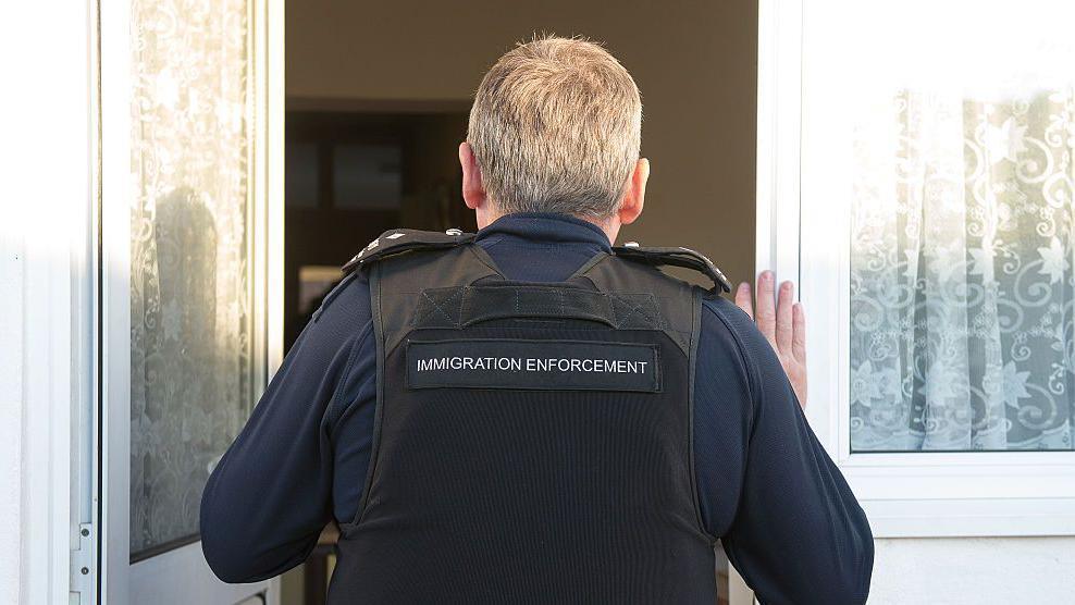 The back of a man with grey hair opens a front door - he is wearing a navy blue long sleeved top and a black vest which reads "Immigration Enforcement"