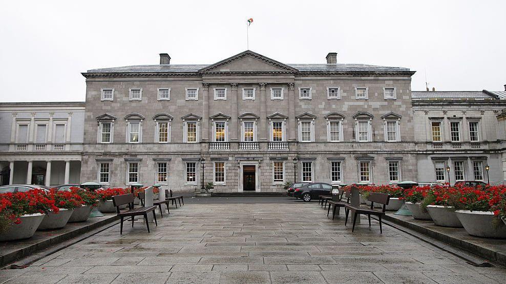 A general view of Leinster House which houses the Seanad chamber, also known as the upper house of the Irish parliament, is pictured in Dublin, Ireland, in 2013. It is a large grey Georgian, Palladian style building with the Irish flag on top. A row of red flowers in pots and seating benches line a walkway leading up to it.
