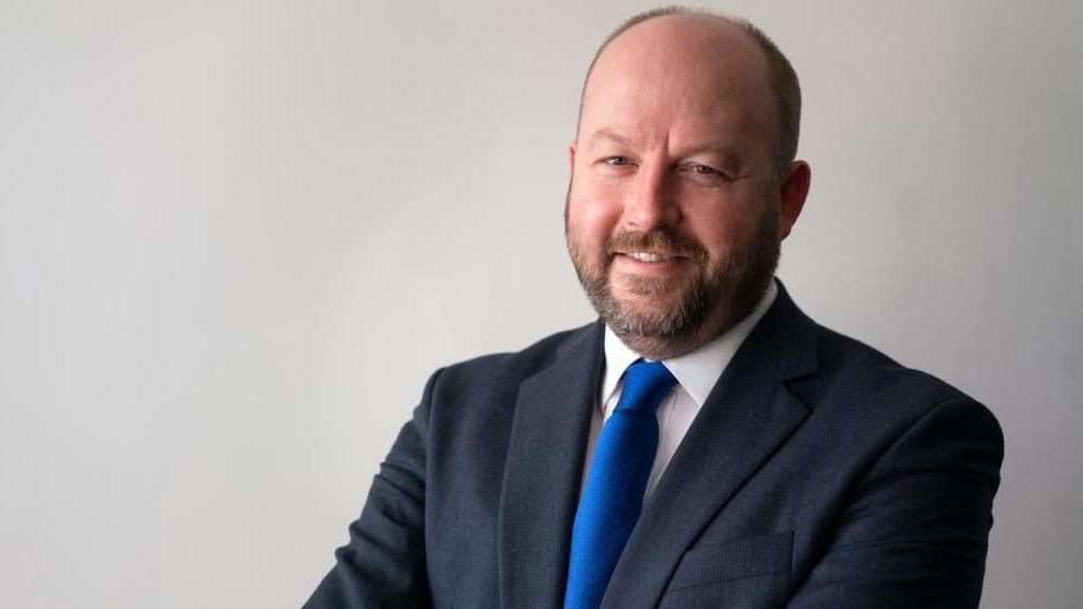 Nick Timothy is  smiling at the camera and is wearing a dark suit jacket over a white shirt with a bright blue tie. He is posed against a plain light-colored background with arms crossed. 