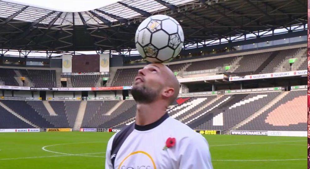 Daniel Cutting with very short hair and dark beard wearing a white Songs of Praise T-shirt and holding a football on his forehead at the MK Dons stadium