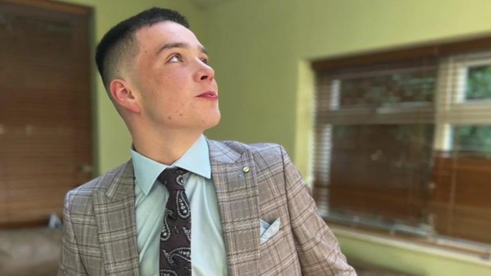 A young boy with dark hair is looking up and to the right. He is wearing a light grey checked suit with a light blue handkerchief in the breast pocket. He wears a light blue shirt and brown paisley patterned tie. In the background are two windows, both with brown blinds pulled down.