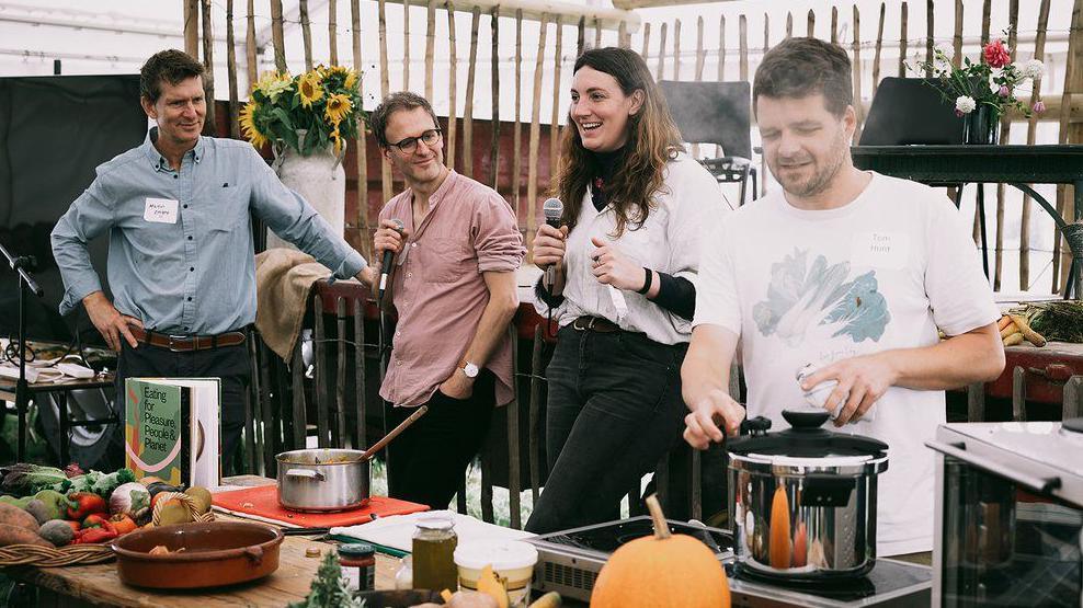 Four people, two holding microphones, talk while another person cooks food in a pot on an electric stove on a table