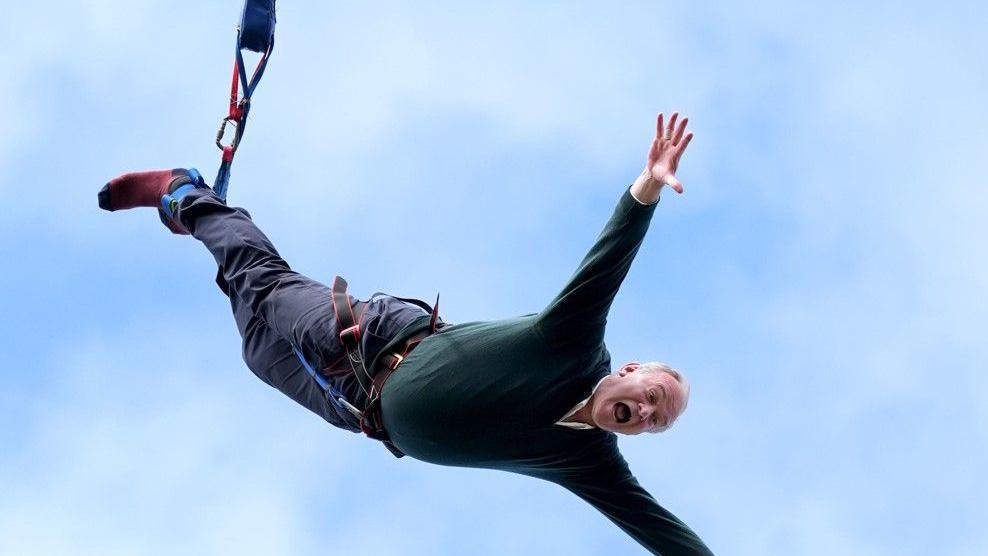 Ed Davey mid bungee jump, with his arms out and a blue sky behind him