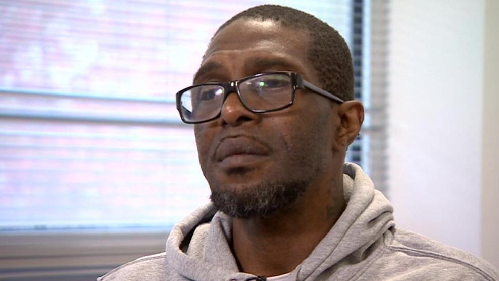 Damion Wright wearing a grey hoodie and thick rectangular glasses with black frames. He has short dark hair, a beard and thin moustache. He has a sad expression on his face. 