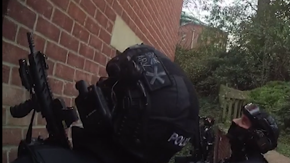 Two firearms officers in uniform with masks covering their heads and mouths. One officer is holding a rifle-like gun and both officers are looking up. 