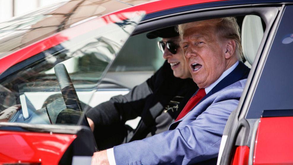 Donald Trump and Elon Musk sit inside a red Tesla; Musk smiles and looks ahead while Trump speaks