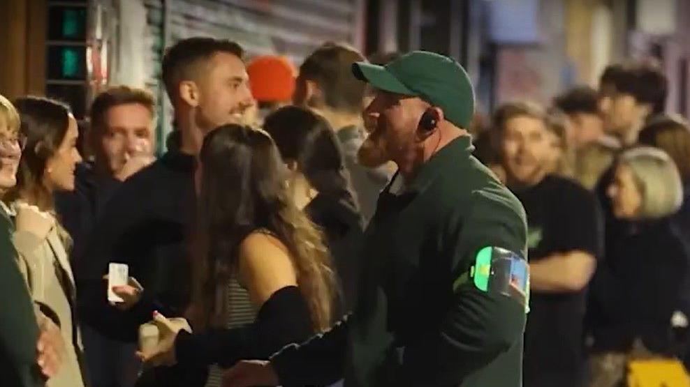 Image shows people queuing for a club in Manchester and a member of a door staff team talking to some of the people in the line