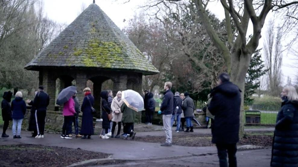 Men and women, some with umbrellas, gather around a stone bandstand with a pointed, tiled roof, on a grey day in Pickering Park, Hull.ull's Pickering Park