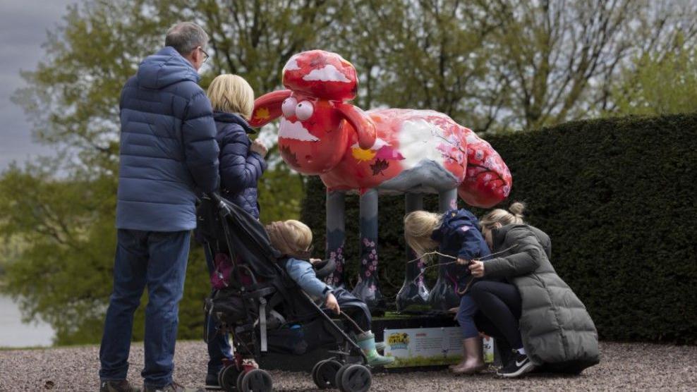 A family is looking at a painted model of a large sheep. There is a man and woman in the image and three children, one of whom is in a pushchair. The woman is crouching down and showing a young girl the details of the artist which are printed on a plinth