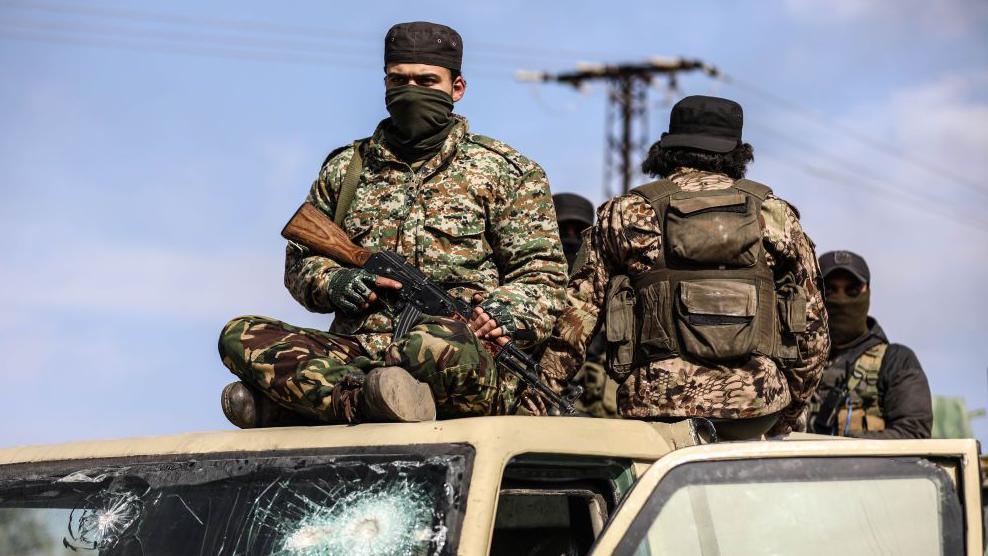 Two soldiers sitting on the roof of a vehicle. One - who is sitting crossed legged and facing the front - is wearing a face covering and is holding a gun. The other has his back to the camera, and is facing two other soldiers who are standing in the back of the vehicle.