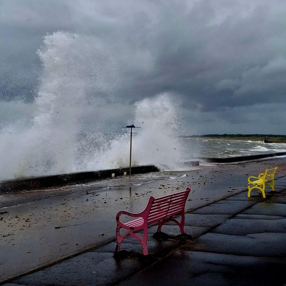 Tall waves crash against the barriers at Prestwick Beach. It appears to be a windy day with dark storm clouds. Two benches, one red and one yellow, sit back from the water.