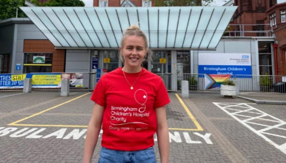 Hannah Hampton in a red t-shirt and blue jeans outside Birmingham Children's Hospital. Her blonde hair is tied back. The hospital's main entrance, signs and ambulance parking bay can be seen behind Hannah.