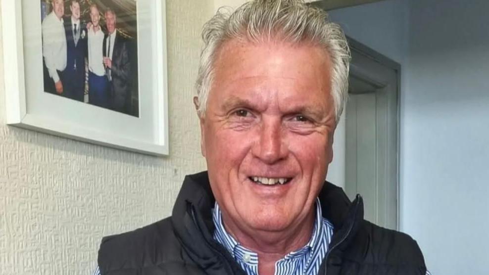 David Moss wearing a pinstriped shirt and gillet. He has grey hair and is smiling at the camera. Behind him on the wall is a wedding photo.