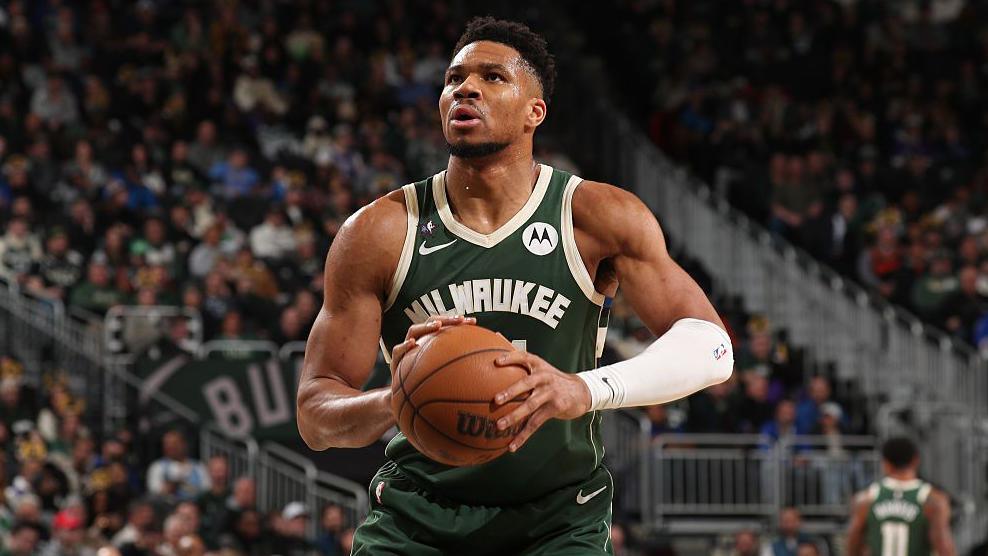 Giannis Antetokounmpo is wearing a green Milwaukee Bucks vest and holding a basketball