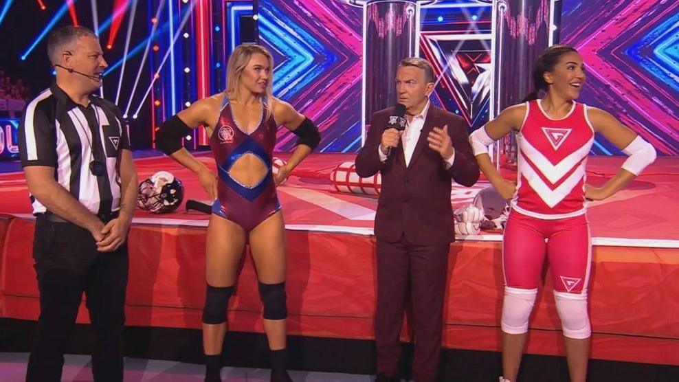 A picture of four people standing on set of the BBC show Gladiators. A referee is on the far left, next to a blonde woman dressed in a red, shiny bodysuit. A woman in a red vest and knee-length leggings is on the far right and Bradley Walsh, in a burgundy suit, is in between the two women. There are big, red pads on the floor behind them that come up to their waist. 