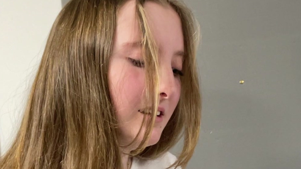 A young woman with fair hair, which is falling in front of her face, singing. She is in front of a neutral background.