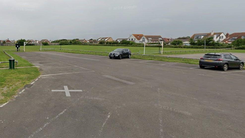A car park next to a large green park. There is one car parked at the site.
