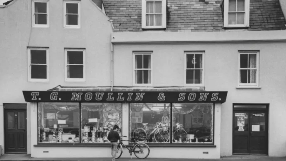 A black and white wide shot of the bicycle shop, T. G. Moullin and Sons. It has a large sign with the shop's name across it, and the front window is filled with bicycles.