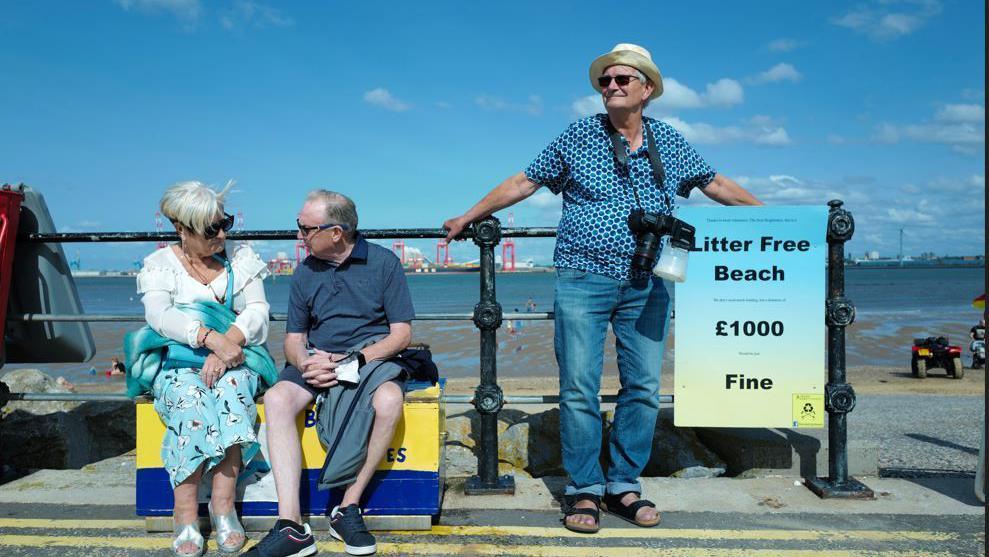 An older man wearing sunglasses, a summer shirt, hat and jeans and sandals, leans on a black fence on a promenade on a sunny day, next to an older couple. A camera is hanging around his neck 