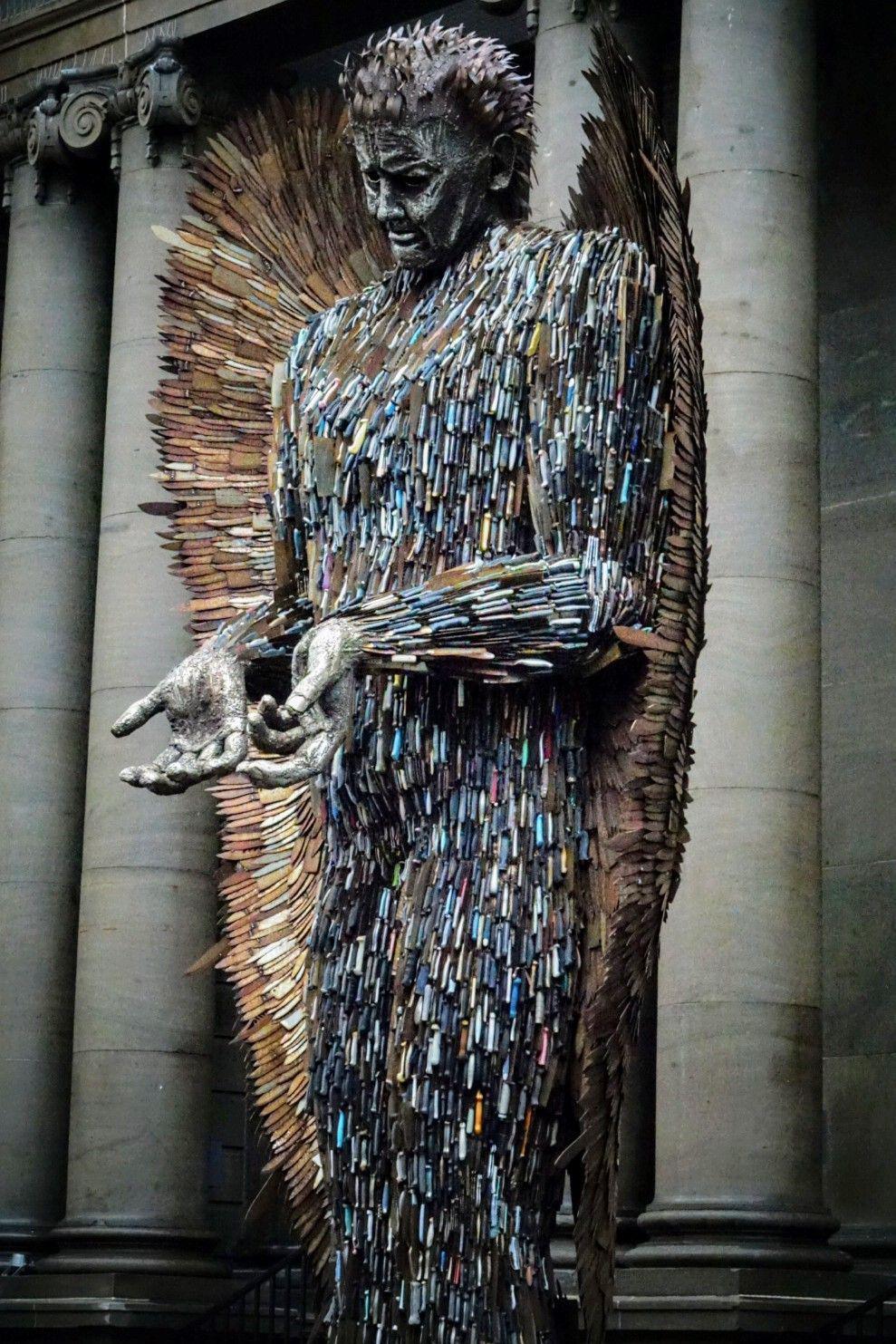 An angel statue completely made of metal knives. The angel is looking down and is holding its hands out.