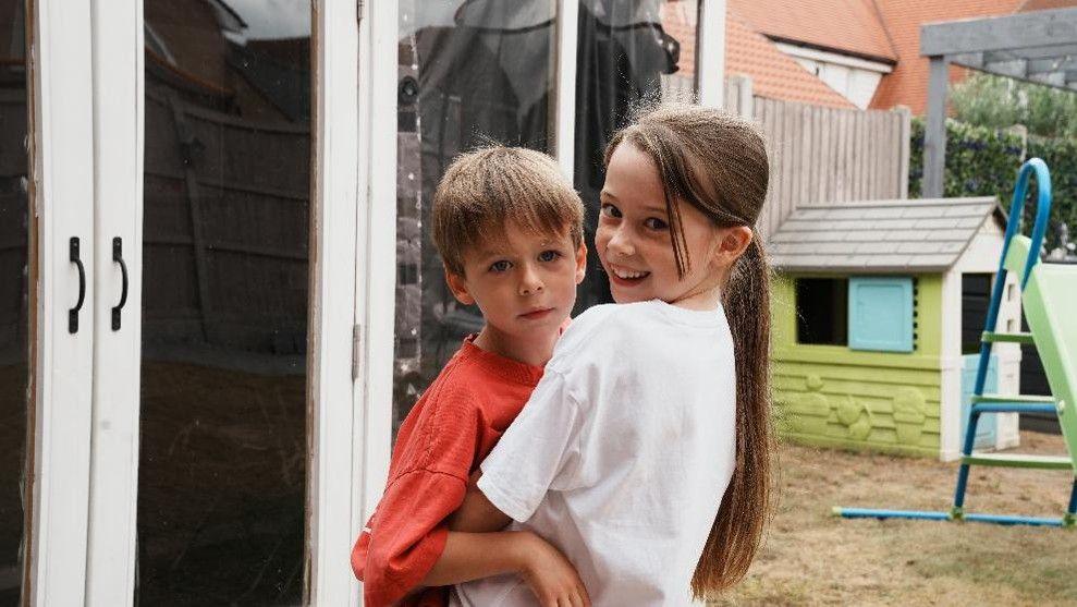 Freddie being embraced by his sister while she turns to smile to the camera. There is a Wendy house and a slide in the background.