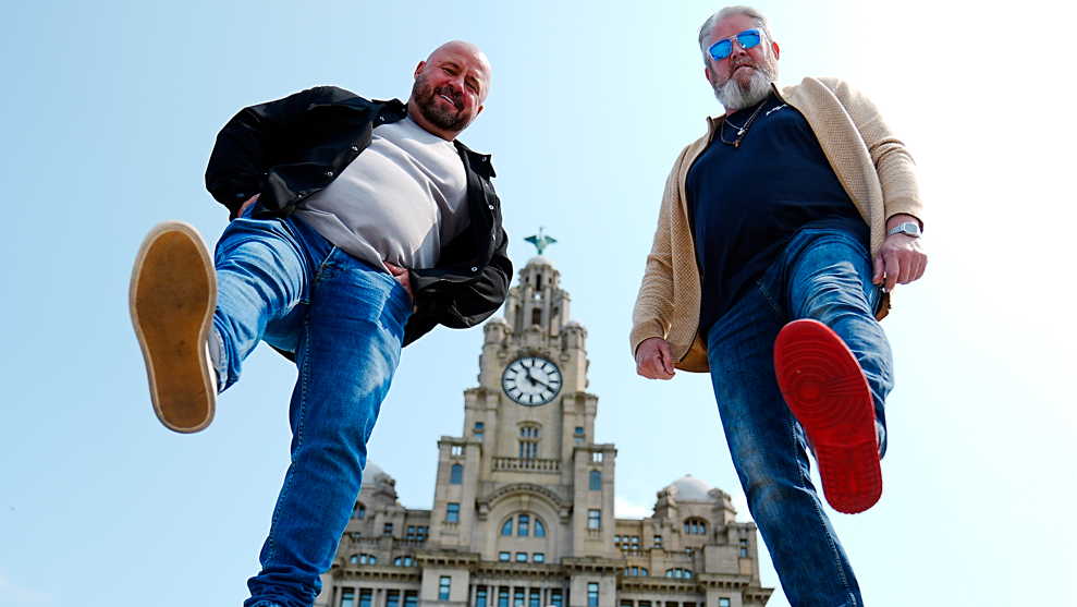 Comedian John May and Tim Edwards pose for a photo with one foot each in the air