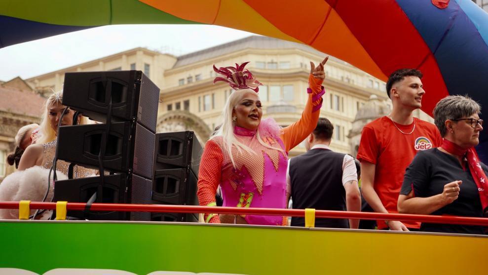 A drag queen who has blonde hair and is wearing a pink and orange outfit. They are standing on top of a an open top bus that is part of a Pride parade. 