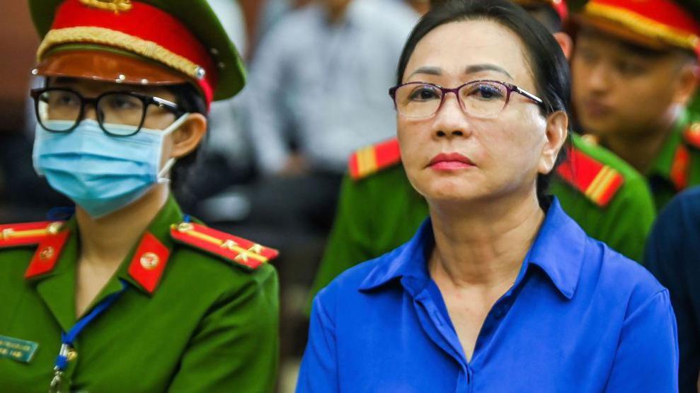 Truong My Lan in a dark blue shirt in court, sitting next to a security officer wearing a mask. 