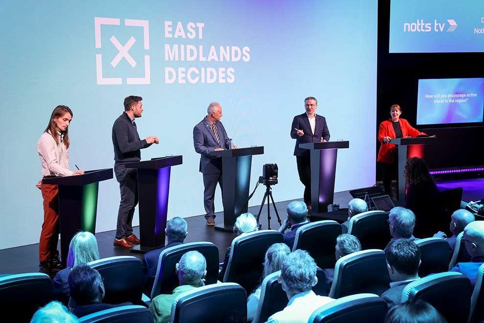 A mayoral debate taking place in Nottingham. Four candidates stand on a stage in front of a large, light-blue screen which reads 'EAST MIDLANDS DECIDES'.