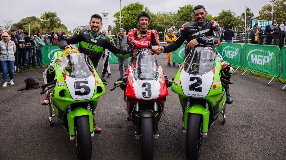 Mike Browne on his red Classic Superbike shaking hands with Rob Hodson and Derek Shiels, who are both sitting on Classic Superbikes that are green.