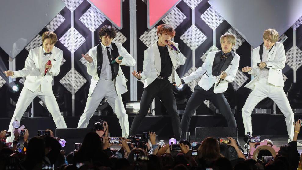 V, Suga, Jin, Jungkook, RM, Jimin, and J-Hope of BTS, dressed in white jackets and black-and-white pants, perform on stage with a mic in each of their hands