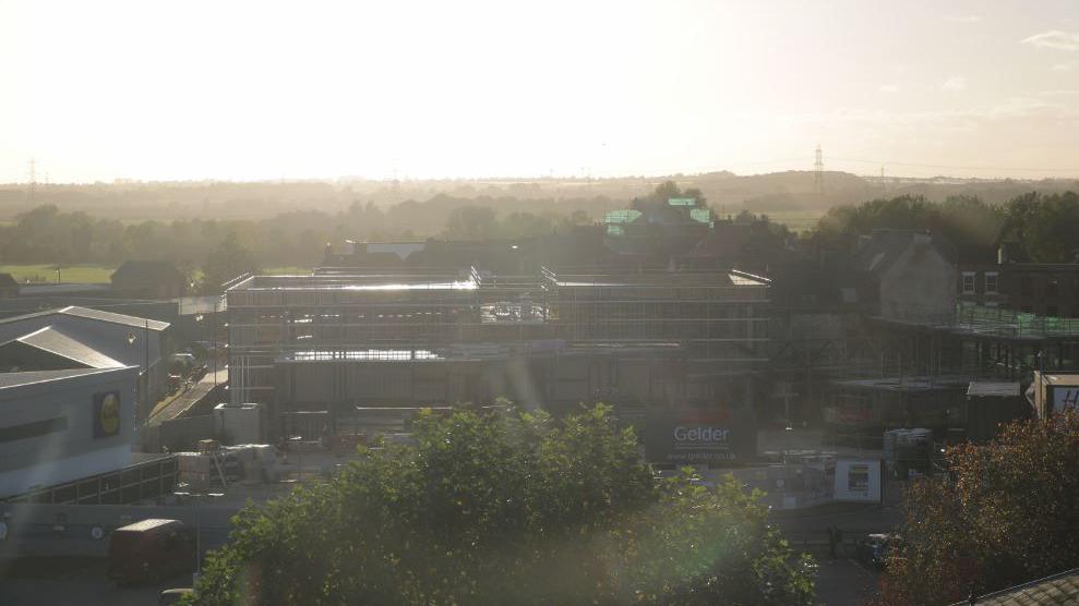 A landscape view of the external structure of a cinema with the word 'Gelder' in the corner, next to a Lidl and other buildings with a tree in the foreground.