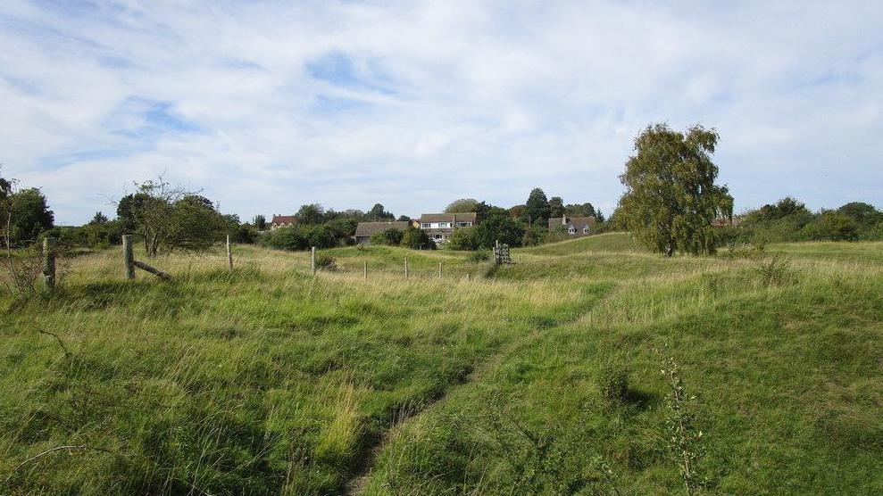 Gently undulating grassy field with trees and houses in the distance, Barnack