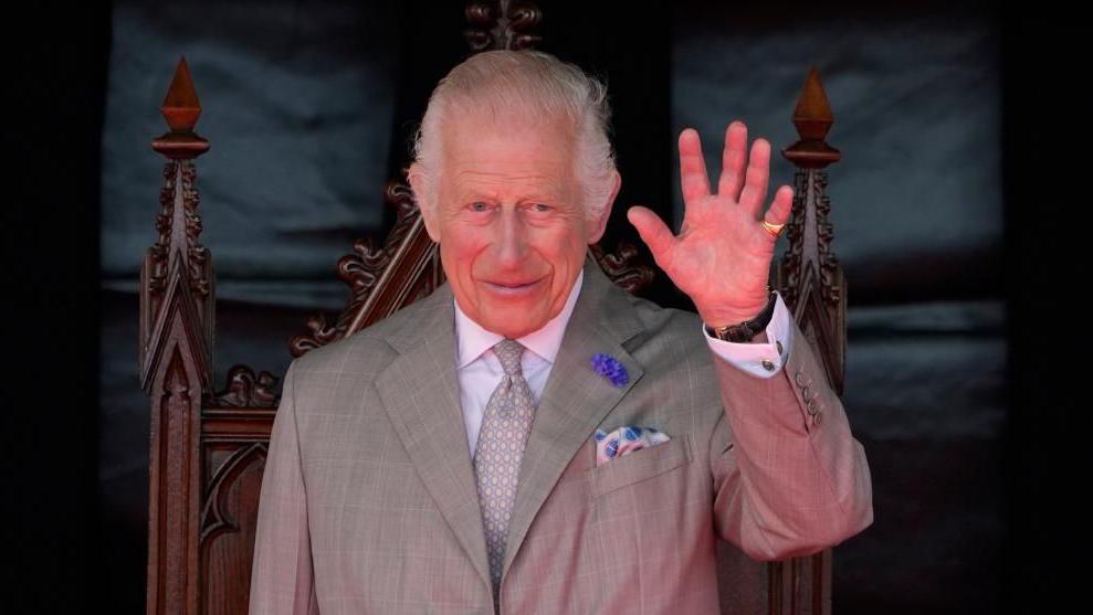 A picture of King Charles. He is waving and wearing a grey suit with blue accents and is stood in front of a wooden chair.