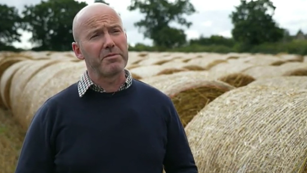 A clean-shaven man in a blue jumper with a checked shirt underneath stood next to hay bales in a field