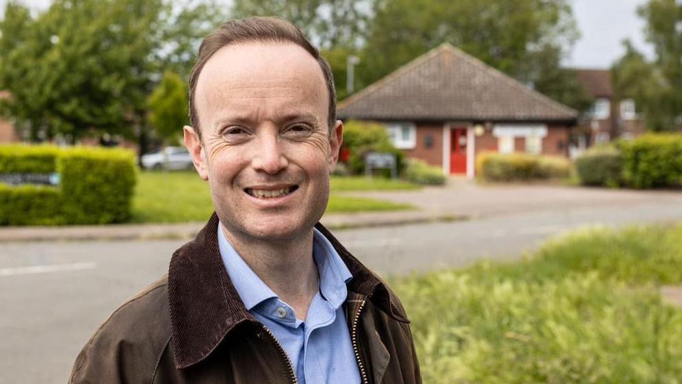 Richard Rout smiles at the camera. He has short brown hair and wears a brown jacket with a blue shirt underneath. He is standing in front of a patch of grass with a bungalow behind him. 