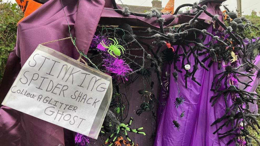 A homemade halloween den which is a tent made from purple fabric which is covered in toy spiders and a sign reading 'stinking spider shack'