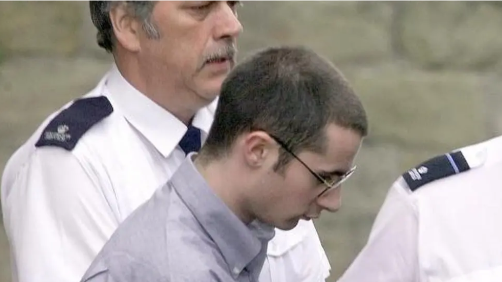 McKilligan being escorted by two police officers in white shirts. McKilligan has short brown hair, is wearing glasses and a blue shirt.