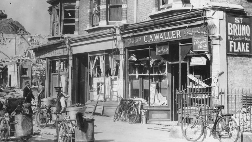 Archive photograph of 1940s London. There are people in the street and several bicycles in front of a row of shops. 