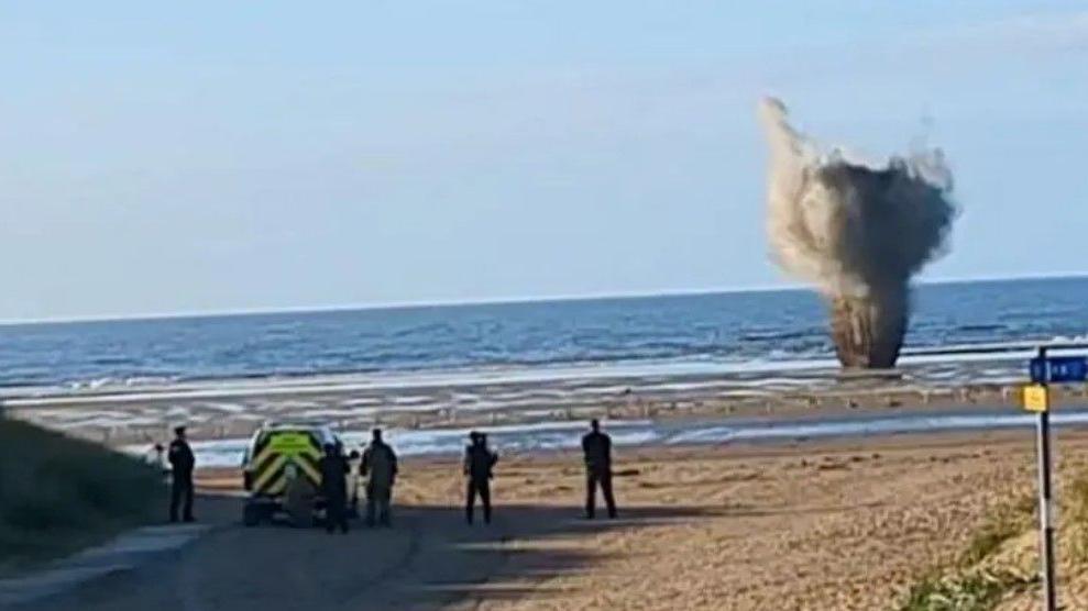 A wide picture of Ainsdale Beach as the WW2 bomb is exploded underwater. Smoke from the detonation can be see above the water, and officers are standing to watch.