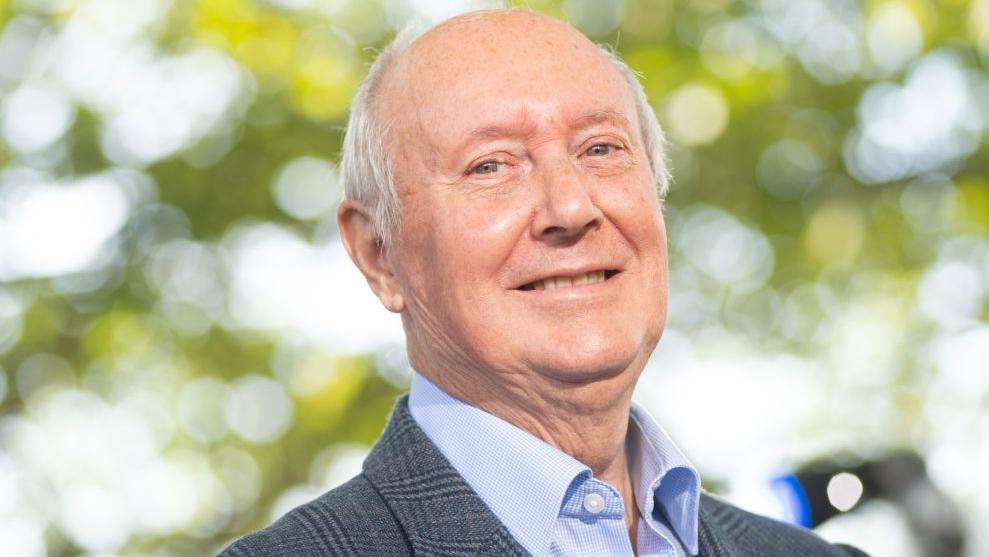 Sir Kenneth Calman in open necked blue shirt and blue checked jacket is smiling  in front of a de-focussed background of trees and sky.