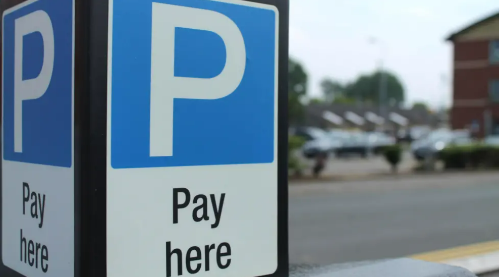 A blue sign with a blue box at the top with a large white 'P'. Underneath is a white block which has the words 'Pay here'. In the background is an out-of-vision parking area.