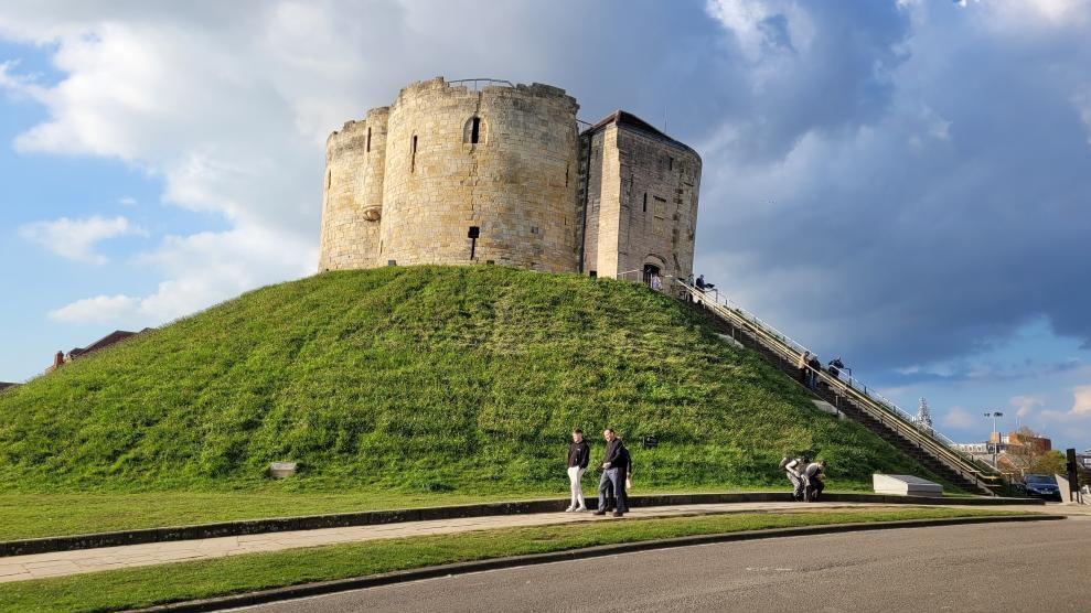 Clifford's Tower in York, on a large grass mound, with steps leading up to the entrance and visitors walking around the landmark.