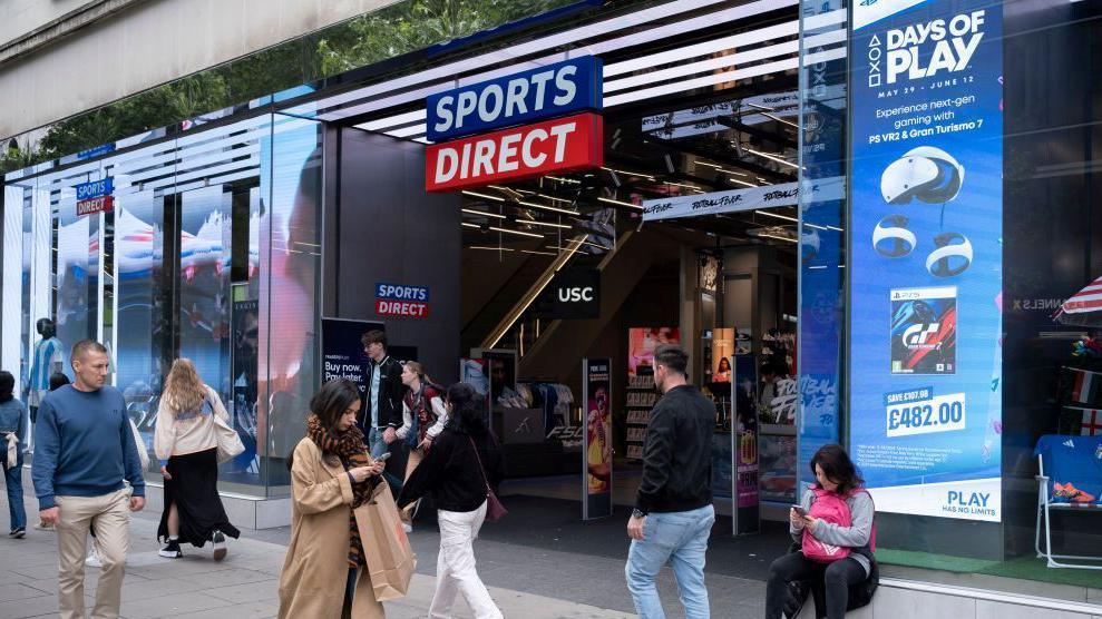 Shoppers and visitors on Oxford Street outside Sports Direct's flagship store in London in June 2024. People are walking outside the shop and coming out of the entrance and one woman, in a beige coat, is looking at her phone. Another woman is sat on a ledge outside the shop looking at her phone. 