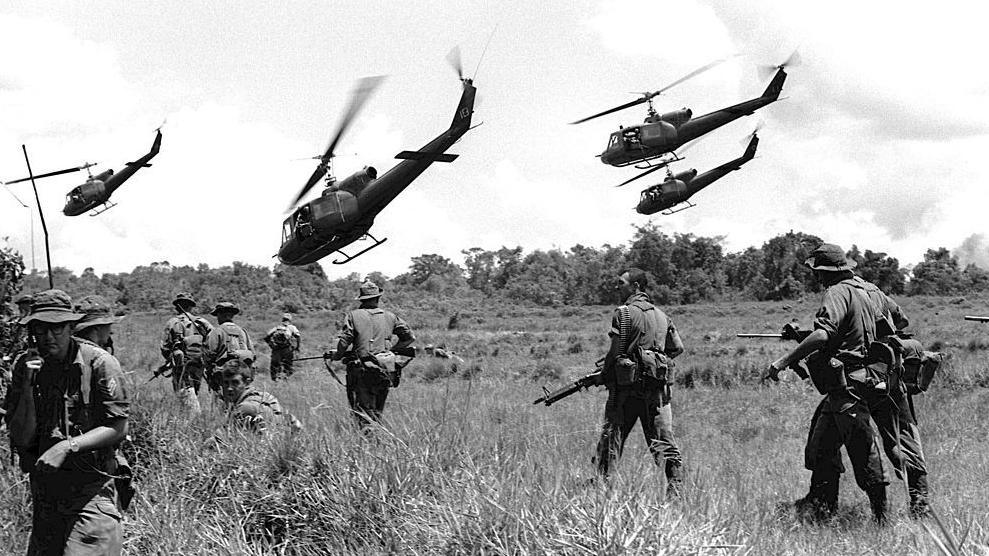 Australian troops carrying guns walk through long grass. Four helicopters fly low over the terrain.