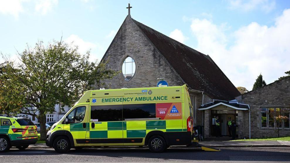 An ambulance parked outside All Saints Church in Swanage - a stone-built building with a cross on the roof and an elliptical window on the gable end